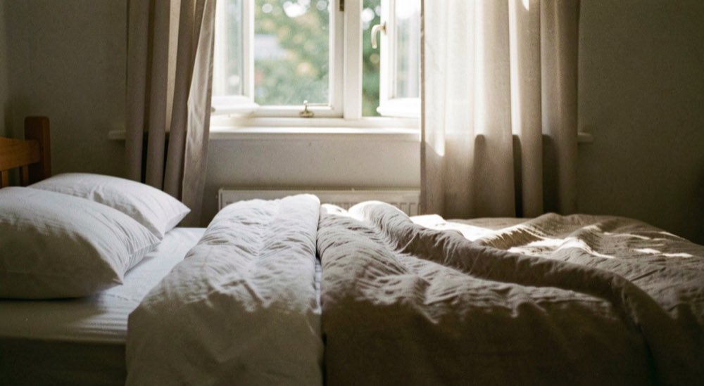 Een opgemaakt bed bij een raam met halfopen gordijnen waar het eerste ochtendlicht doorheen valt.